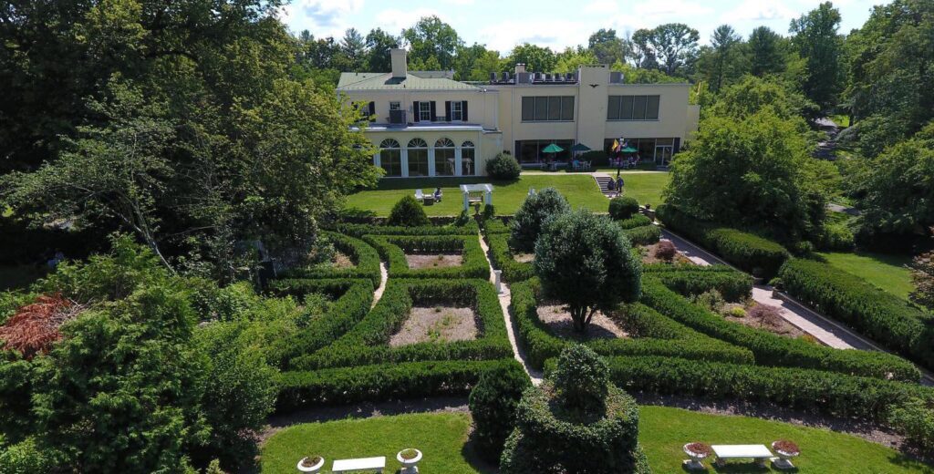 An aerial view of a formal garden with neatly trimmed hedges and pathways, leading up to a large, elegant building surrounded by trees. The garden includes benches and small decorative features, creating a peaceful, well-manicured landscape.