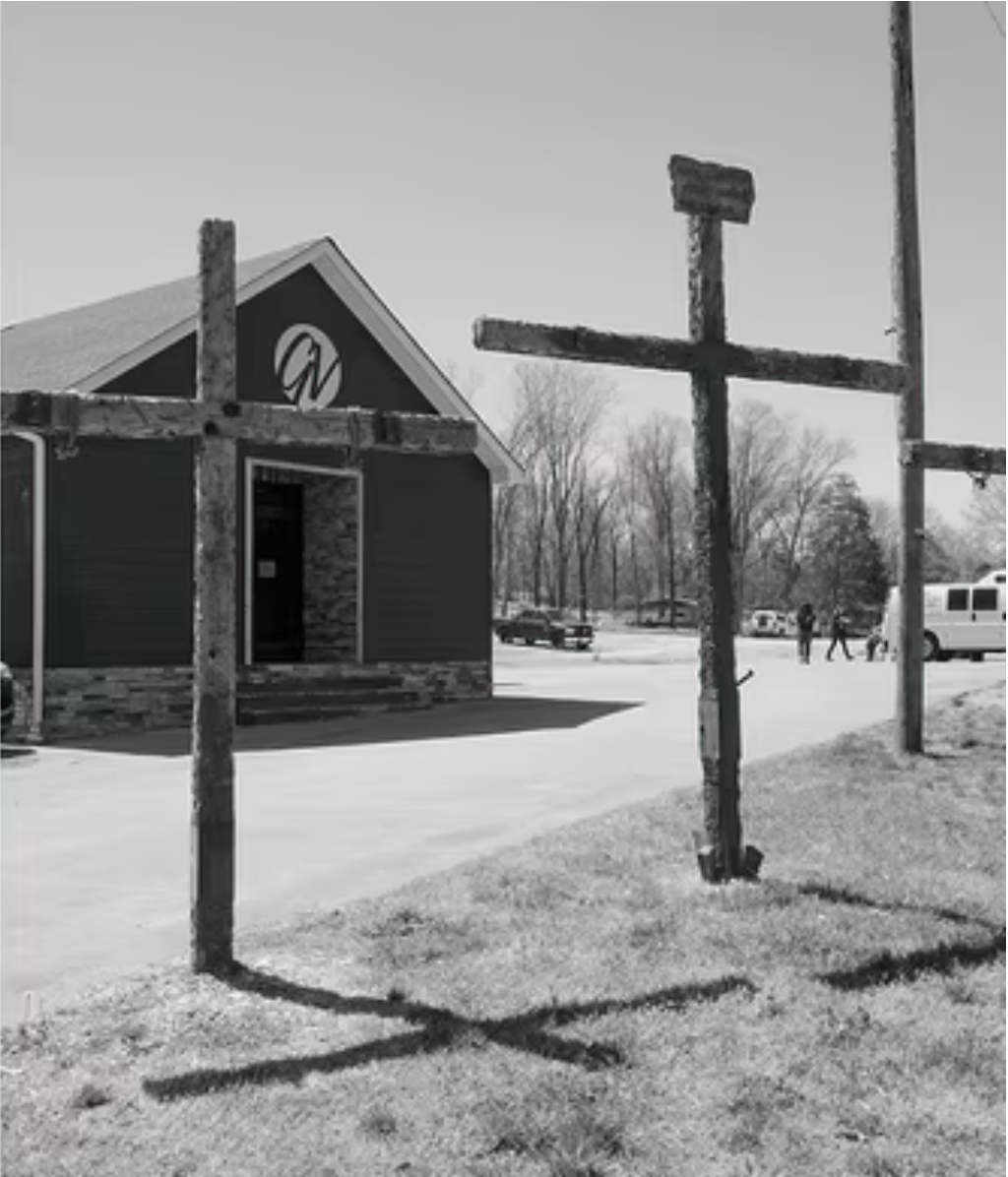 A black-and-white photo showing two large wooden crosses standing in front of a small building, with shadows cast on the grass. The building has a sign with a stylized "W," and a few people and vehicles are visible in the background.