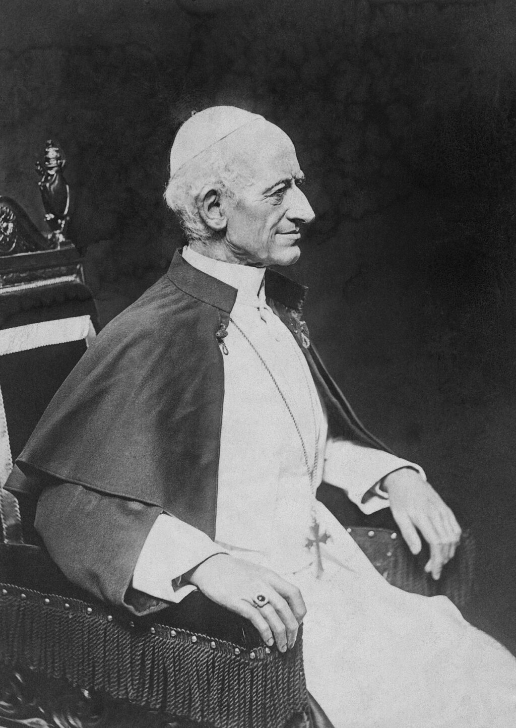 A black-and-white portrait of Pope Leo XIII, seated in profile on a chair, wearing papal vestments including a white cassock and a cape. He has a serene expression, with his hands resting on the arms of the chair, and wears a ring on his right hand.