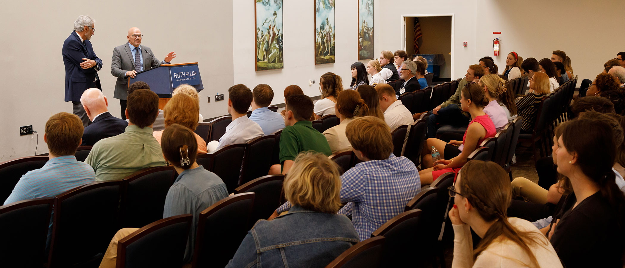 A speaker addresses an audience at a Faith & Law event in Washington, D.C. Two men stand at the front, one speaking at a podium while the audience listens attentively. The room features artwork on the walls, and attendees are seated in rows.