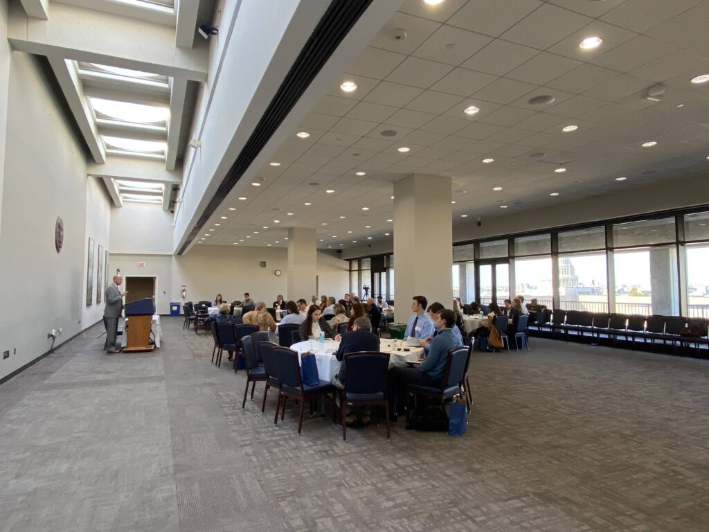 Photo of 2024 conference. Speaker is at podium. Capitol building is visible through windows.